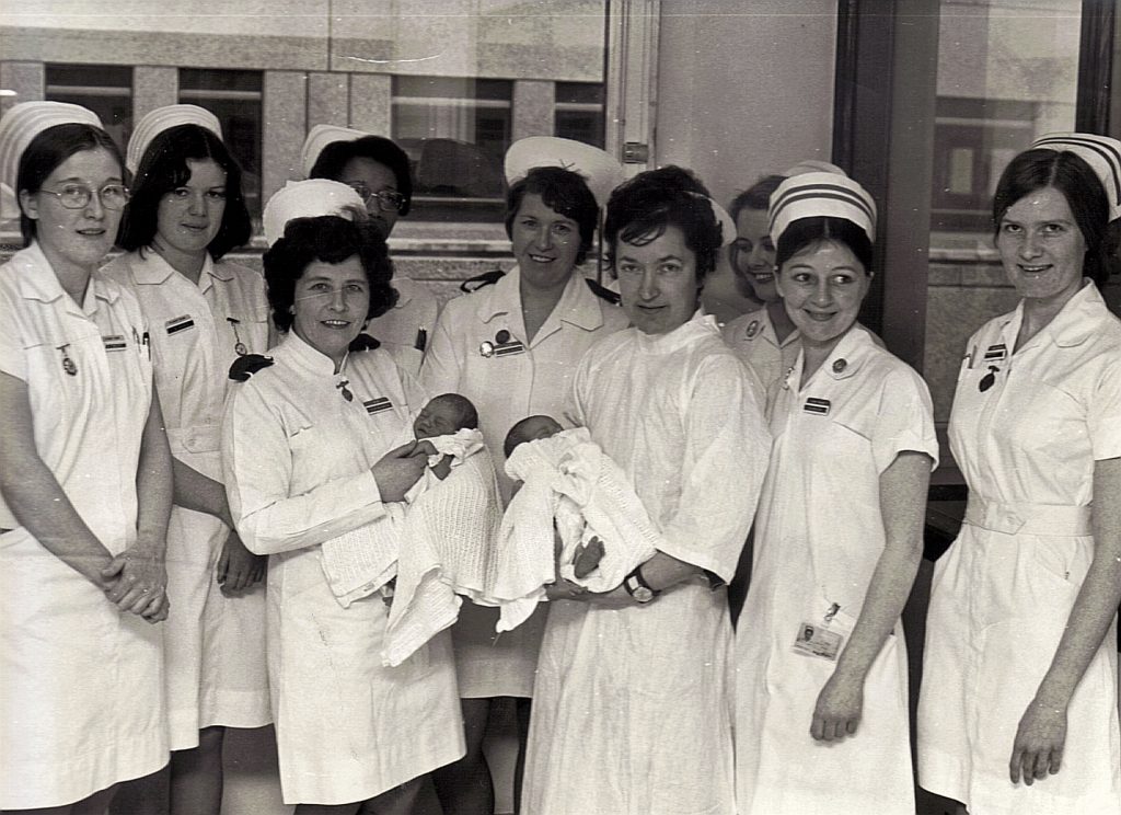 Photograph of 8 nurses in white uniforms and hats posing for the camera. The two midwives in the centre are each holding a new-born baby
