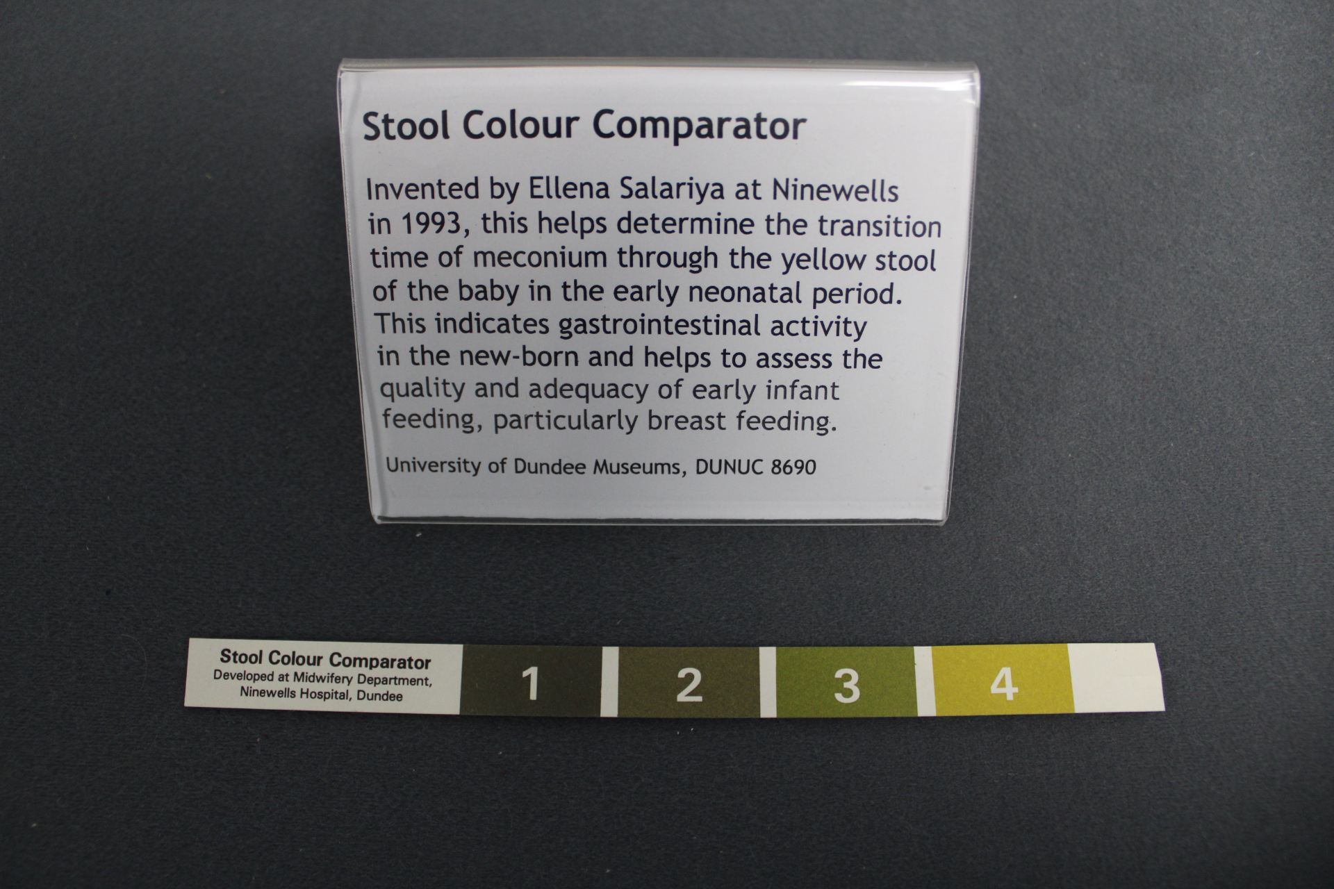 A small strip of paper with 4 shades from dark green to yellow for comparing the colour of a baby's stool. Behind the comparator is a museum label explaining the object. 