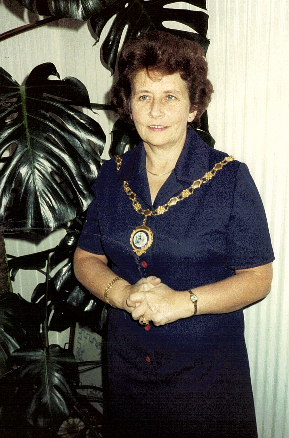 image of Ellena Salariya wearing the Chain of Office for the Royal College of Midwives. She is wearing a navy collared dress and standing in front of a plant with her hands clasped. 