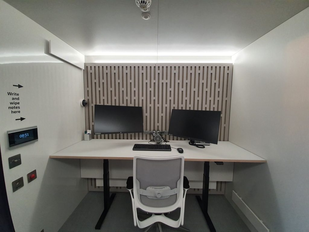 Desk with 2 monitors, keyboard, mouse, keyboard and fan sits in front of a grey acoustic panel wall, CCTV camera, door intercom and emergency alarm buttons on the left and a grey and white chair at the desk
